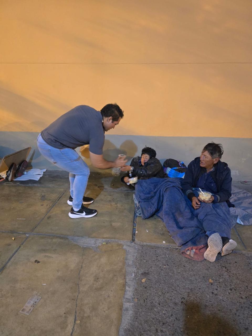 Un voluntario se agacha para entregar un vaso a dos personas que descansan en la vereda de noche