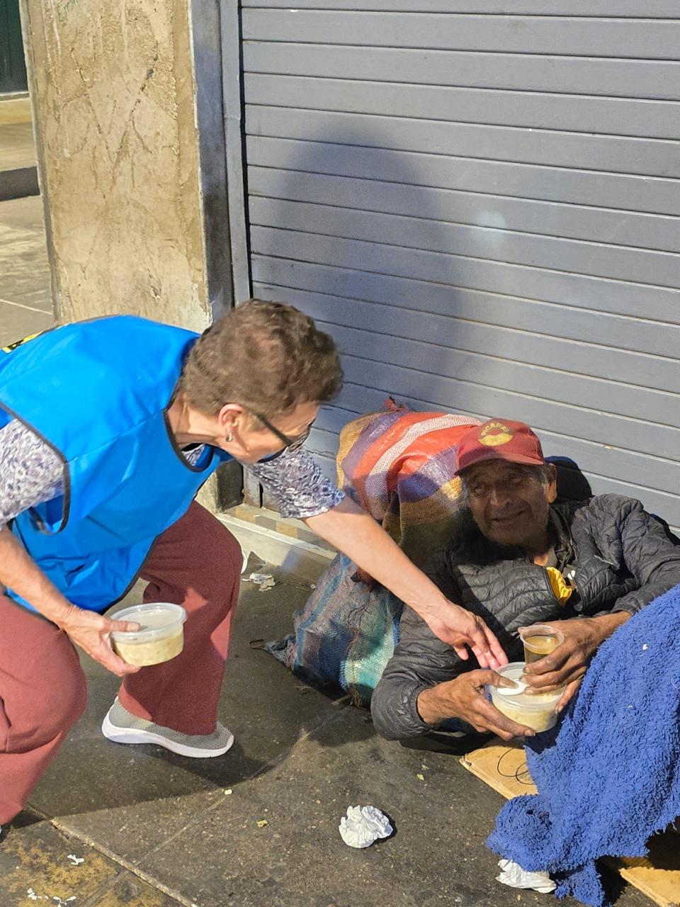 Una voluntaria con chaleco azul entrega una porción de comida a un hombre sentado junto a una cortina metálica