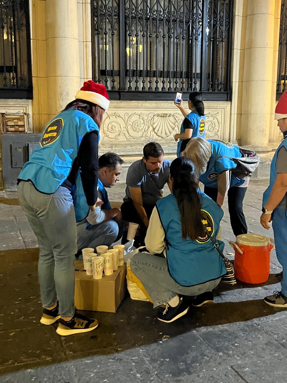 Voluntarios con gorros navideños sirven bebida caliente en vasos sobre una caja de cartón