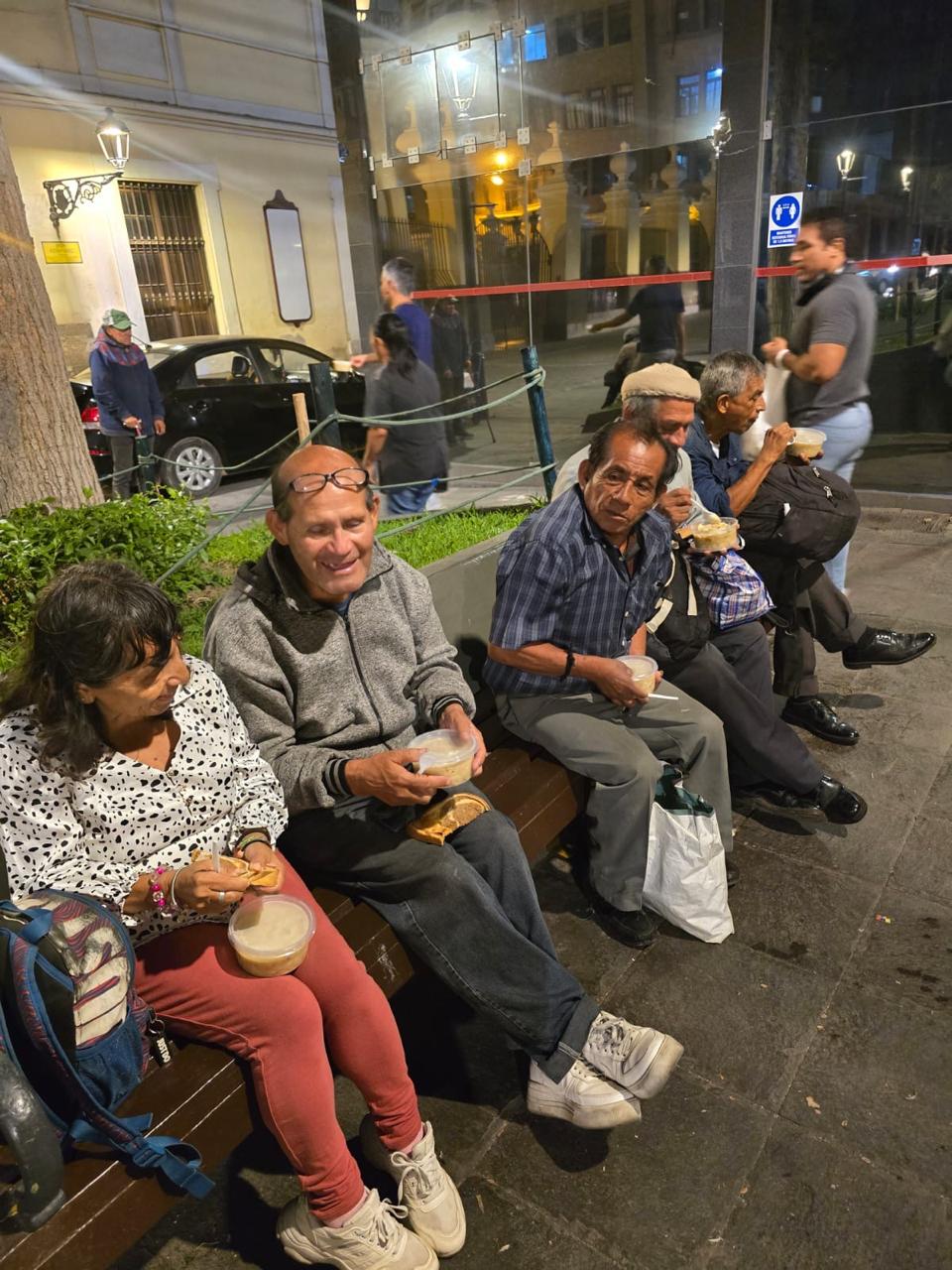 Personas sentadas en una banca de la plaza comiendo la comida que recibieron