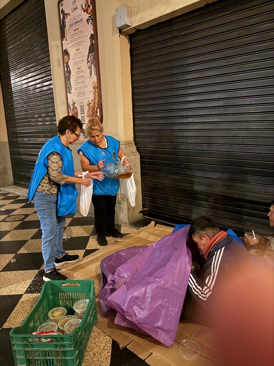 Dos voluntarias sirven bebida y entregan bolsas a una persona que descansa bajo una persiana