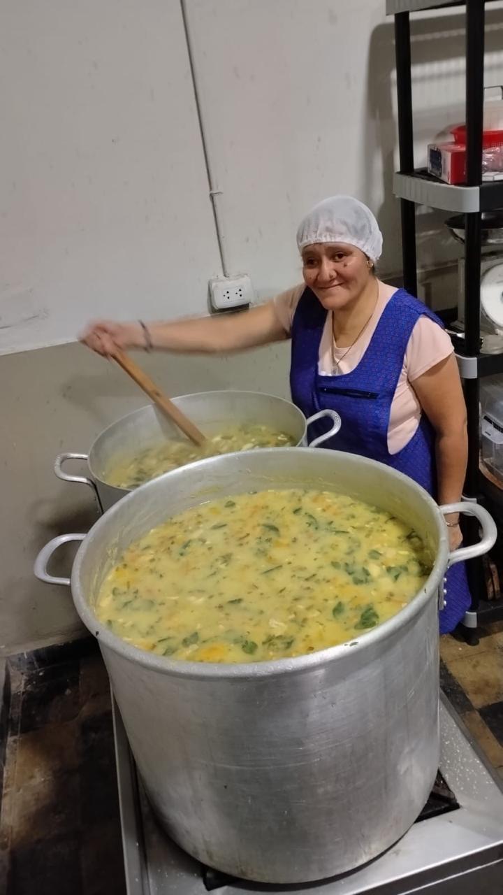 Una voluntaria con gorro y delantal azul sonríe mientras revuelve una olla grande de sopa en la cocina de Misión Compartir