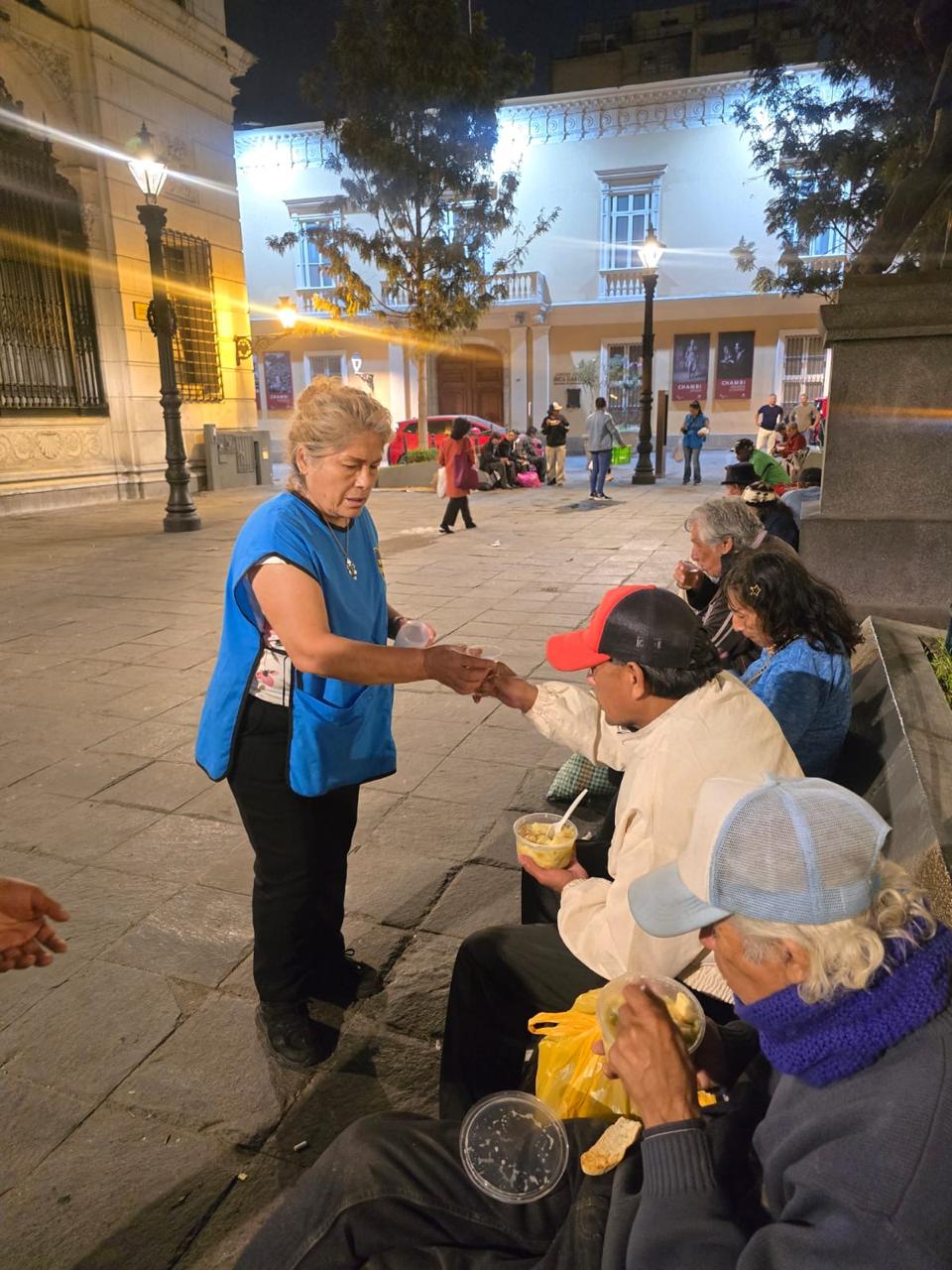 Una voluntaria de Misión Compartir con chaleco azul entrega una porción de comida a una persona sentada en una plaza, de noche