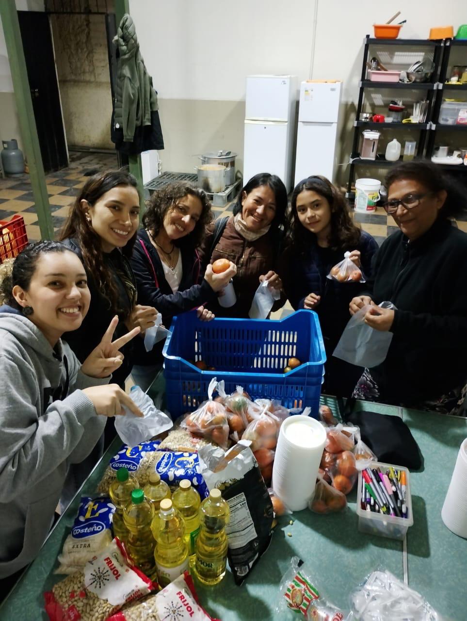 Seis voluntarias sonríen alrededor de una caja con tomates, aceite, menestras y plumones para escribir mensajes en las porciones